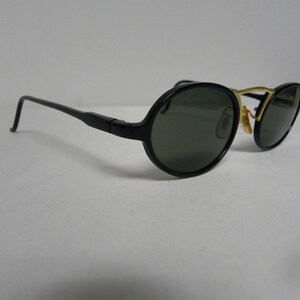 May include: Black sunglasses with oval lenses and a gold-coloured bridge. The frames are black with a sleek, modern design. The lenses are a dark green colour, providing shade and style. The sunglasses are set against a white background.