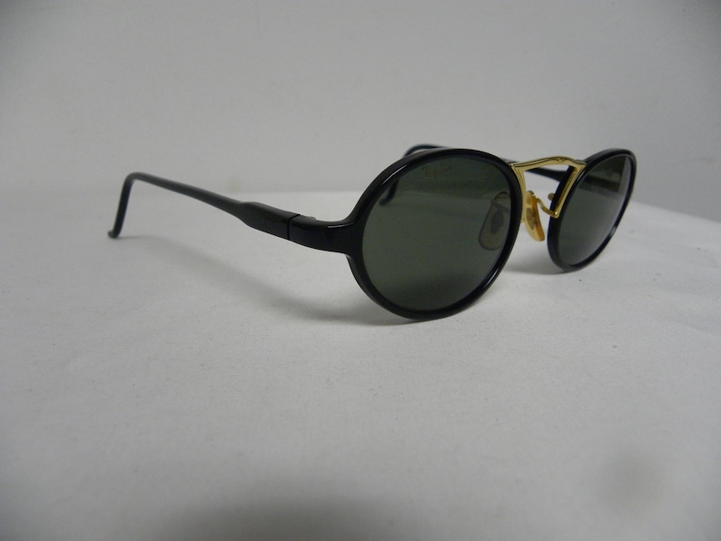 May include: Black sunglasses with oval lenses and a gold-coloured bridge. The frames are black with a sleek, modern design. The lenses are a dark green colour, providing shade and style. The sunglasses are set against a white background.