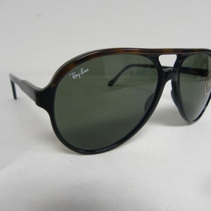 May include: Black and brown aviator sunglasses with green lenses. The sunglasses have a Ray-Ban logo on the right lens.