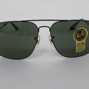 May include: Black Ray-Ban sunglasses with green lenses. The sunglasses have a square frame and a gold sticker on the right lens that says "Ray-Ban" and "G-15 Lens".