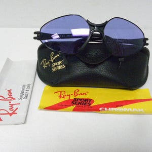 May include: Black Ray-Ban Sport Series sunglasses with purple lenses and a black leather case. The sunglasses have the text "Ray-Ban Sport Series" on the side. The case has the text "Ray-Ban Sport Series" on the front. The sunglasses come with a yellow and red card that says "with CHROMAX".