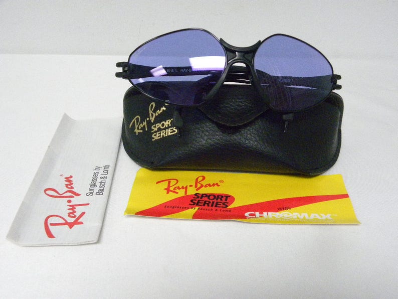 May include: Black Ray-Ban Sport Series sunglasses with purple lenses and a black leather case. The sunglasses have the text "Ray-Ban Sport Series" on the side. The case has the text "Ray-Ban Sport Series" on the front. The sunglasses come with a yellow and red card that says "with CHROMAX".