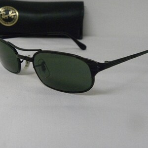 Puede incluir: Gafas de sol negras con lentes verdes y montura de metal. Las gafas tienen un dise&ntilde;o texturizado en las patillas.