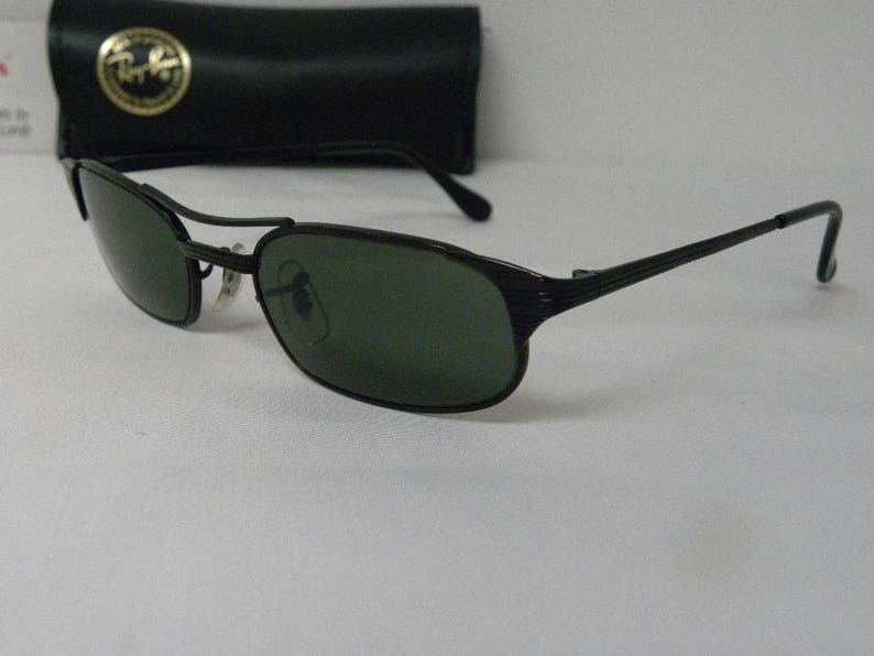 Puede incluir: Gafas de sol negras con lentes verdes y montura de metal. Las gafas tienen un dise&ntilde;o texturizado en las patillas.