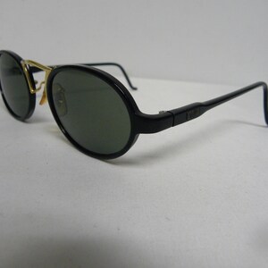 May include: Black oval sunglasses with dark green lenses and gold-coloured accents. The sunglasses have a sleek, modern design with a black frame and temples. The lenses are a dark green colour, providing protection from the sun.