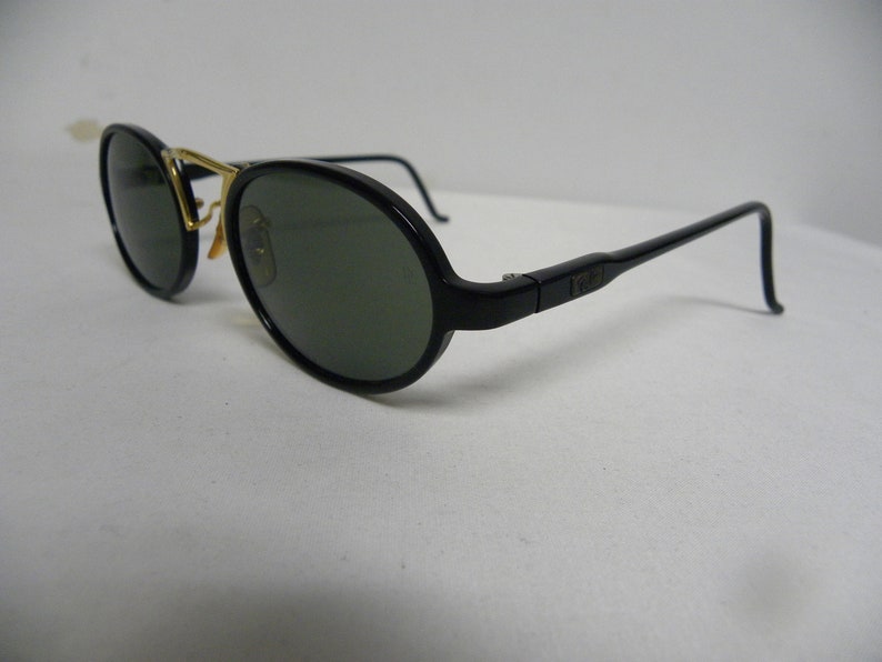 May include: Black oval sunglasses with dark green lenses and gold-coloured accents. The sunglasses have a sleek, modern design with a black frame and temples. The lenses are a dark green colour, providing protection from the sun.