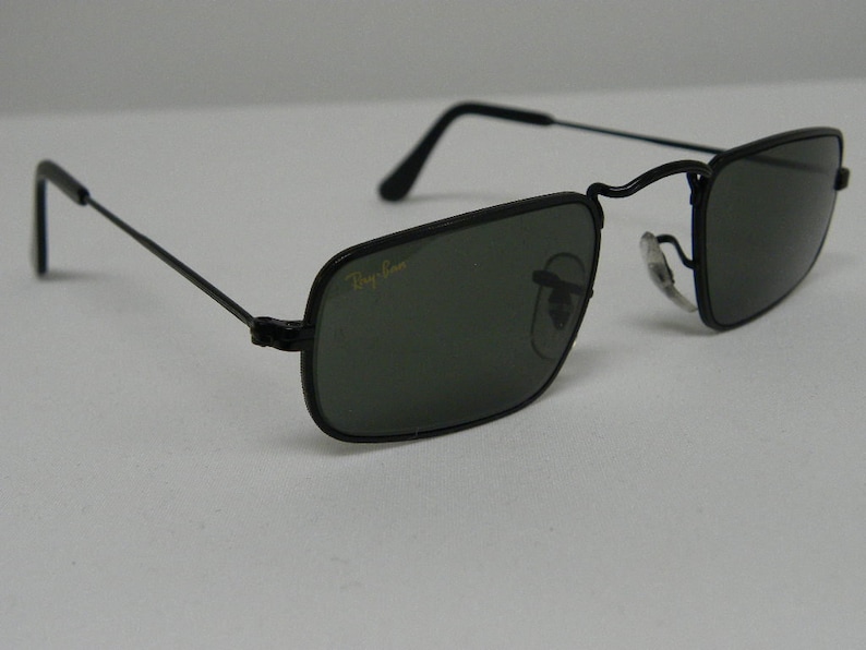 May include: Black square-shaped sunglasses with a thin metal frame and dark green lenses. The sunglasses have the Ray-Ban logo on the frame.