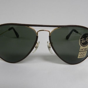 May include: Brown leather aviator sunglasses with gold frames and green lenses. The sunglasses have a Ray-Ban logo on the left lens and a G-15 Lens sticker on the right lens.