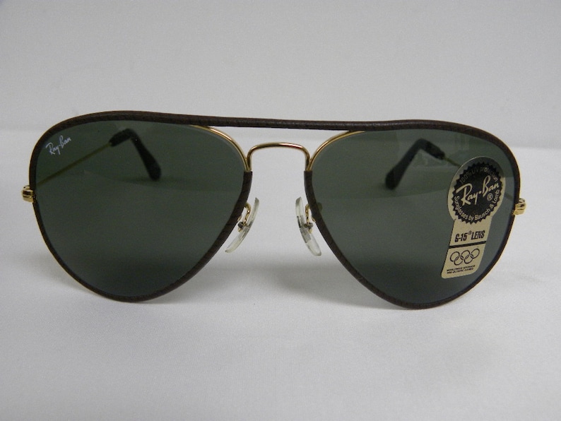 May include: Brown leather aviator sunglasses with gold frames and green lenses. The sunglasses have a Ray-Ban logo on the left lens and a G-15 Lens sticker on the right lens.