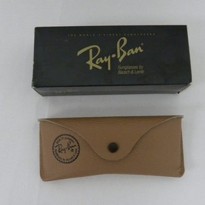 May include: A black Ray-Ban sunglasses box with gold lettering that reads "Ray-Ban Sunglasses by Bausch & Lomb". The box is next to a brown leather case with a Ray-Ban logo embossed on the front.