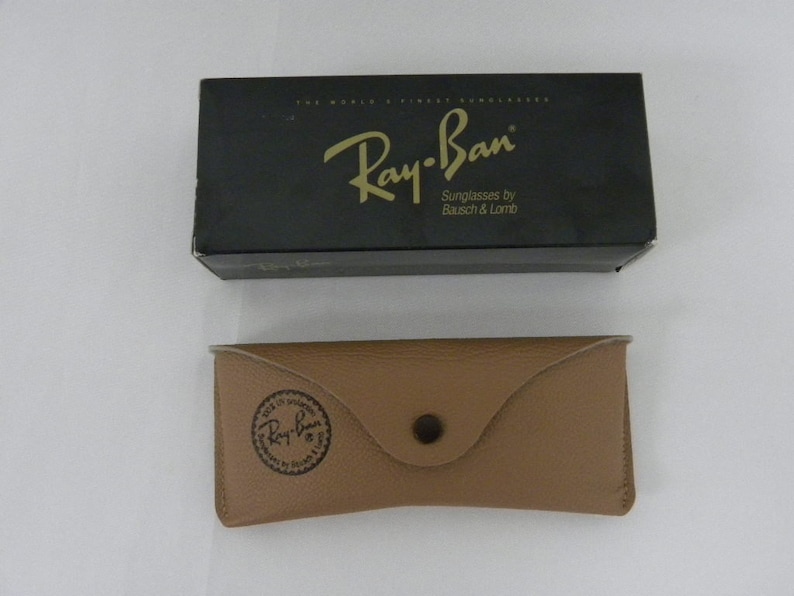 May include: A black Ray-Ban sunglasses box with gold lettering that reads "Ray-Ban Sunglasses by Bausch & Lomb". The box is next to a brown leather case with a Ray-Ban logo embossed on the front.
