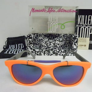 May include: Orange sunglasses with purple lenses and a gray frame. The sunglasses are in a box with the words "Killer Loop" and "Moments before detonation" on it.