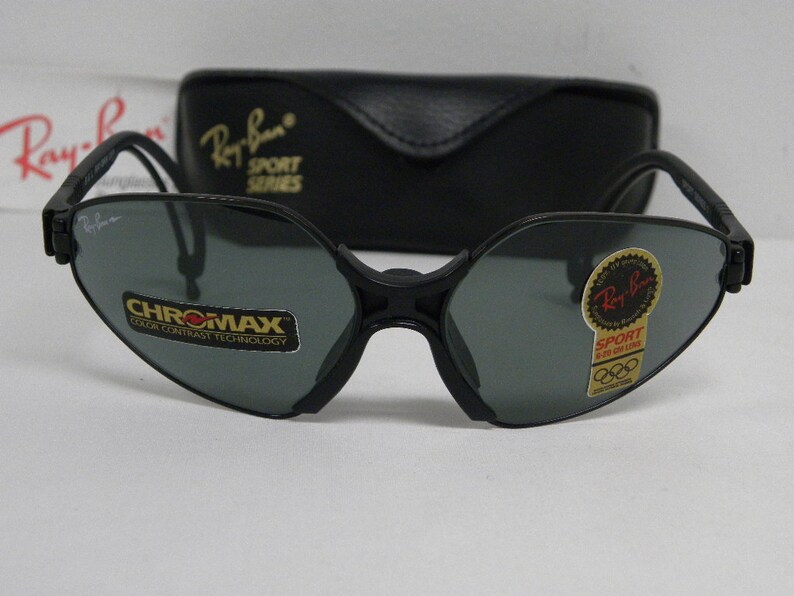 May include: Black Ray-Ban Sport Series sunglasses with gray lenses. The sunglasses have a unique, angular shape and feature the Ray-Ban logo on the side. The lenses have a "Chromax" label, indicating they have color contrast technology. The sunglasses come with a black case.