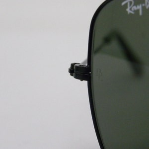 May include: Close-up of a black Ray-Ban sunglasses frame with green lenses. The Ray-Ban logo is visible on the frame. The sunglasses have a metal hinge with a small metal piece that says "RB".