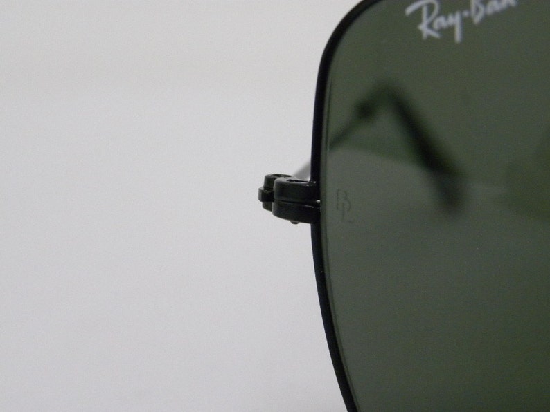 May include: Close-up of a black Ray-Ban sunglasses frame with green lenses. The Ray-Ban logo is visible on the frame. The sunglasses have a metal hinge with a small metal piece that says "RB".
