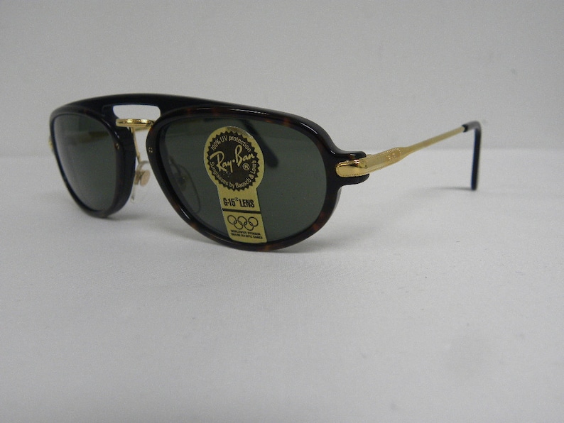 May include: Ray-Ban sunglasses with a dark tortoise shell frame and gold-tone accents. The lenses are dark green, and the sunglasses have a classic aviator shape. A sticker on the lens reads "Ray-Ban G-15 LENS".