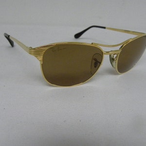 May include: Gold-tone Ray-Ban sunglasses with brown lenses. The sunglasses have a classic aviator style with a metal frame.