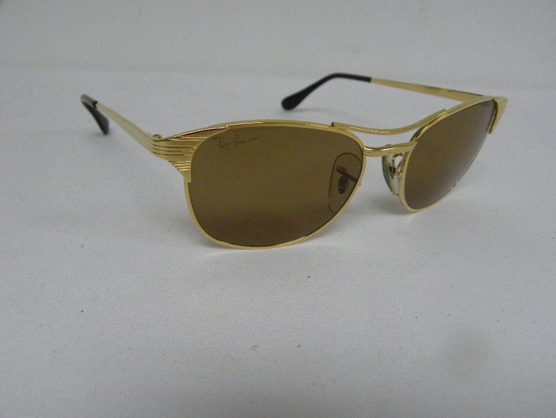 May include: Gold-tone Ray-Ban sunglasses with brown lenses. The sunglasses have a classic aviator style with a metal frame.