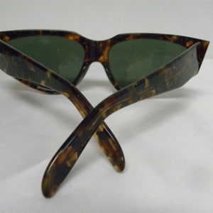 K&ouml;nnte beinhalten: Schildkr&ouml;ten-Sonnenbrille mit gr&uuml;nen Gl&auml;sern. Die Sonnenbrille ist in der Mitte gefaltet, die B&uuml;gel sind gekreuzt.