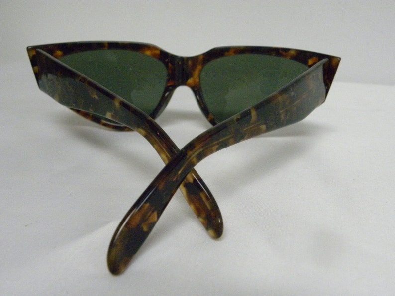 K&ouml;nnte beinhalten: Schildkr&ouml;ten-Sonnenbrille mit gr&uuml;nen Gl&auml;sern. Die Sonnenbrille ist in der Mitte gefaltet, die B&uuml;gel sind gekreuzt.