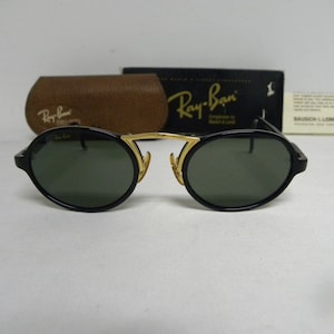 May include: Black oval Ray-Ban sunglasses with gold detailing. The sunglasses feature dark green lenses and are presented with a brown leather case, a black box, and a white tag. The Ray-Ban logo is visible on the sunglasses and case.