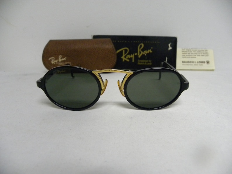May include: Black oval Ray-Ban sunglasses with gold detailing. The sunglasses feature dark green lenses and are presented with a brown leather case, a black box, and a white tag. The Ray-Ban logo is visible on the sunglasses and case.
