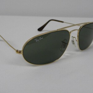 May include: Gold-framed aviator sunglasses with dark green lenses. The Ray-Ban logo is visible on the lens. The sunglasses have a double bridge and thin temples with black tips. The glasses are set against a white background.