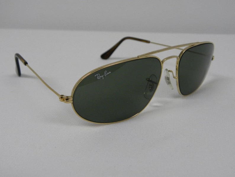 May include: Gold-framed aviator sunglasses with dark green lenses. The Ray-Ban logo is visible on the lens. The sunglasses have a double bridge and thin temples with black tips. The glasses are set against a white background.
