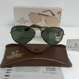 May include: Ray-Ban Leathers aviator sunglasses with green lenses and a brown leather case. The sunglasses have a gold frame and a gold Ray-Ban logo on the right lens. The case has the Ray-Ban Leathers logo embossed on the front.