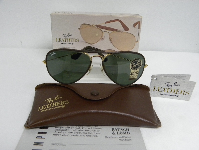 May include: Ray-Ban Leathers aviator sunglasses with green lenses and a brown leather case. The sunglasses have a gold frame and a gold Ray-Ban logo on the right lens. The case has the Ray-Ban Leathers logo embossed on the front.