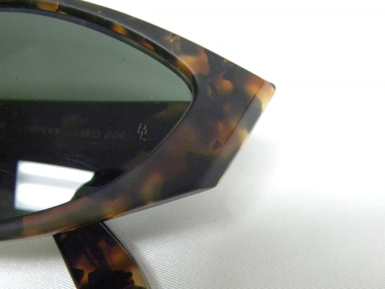 K&ouml;nnte beinhalten: Nahaufnahme einer Sonnenbrille aus Schildpatt mit gr&uuml;nen Gl&auml;sern. Die Sonnenbrille hat eine einzigartige, eckige Form und der Text "Ray-Ban 54 024/3M" ist auf dem Gestell sichtbar.
