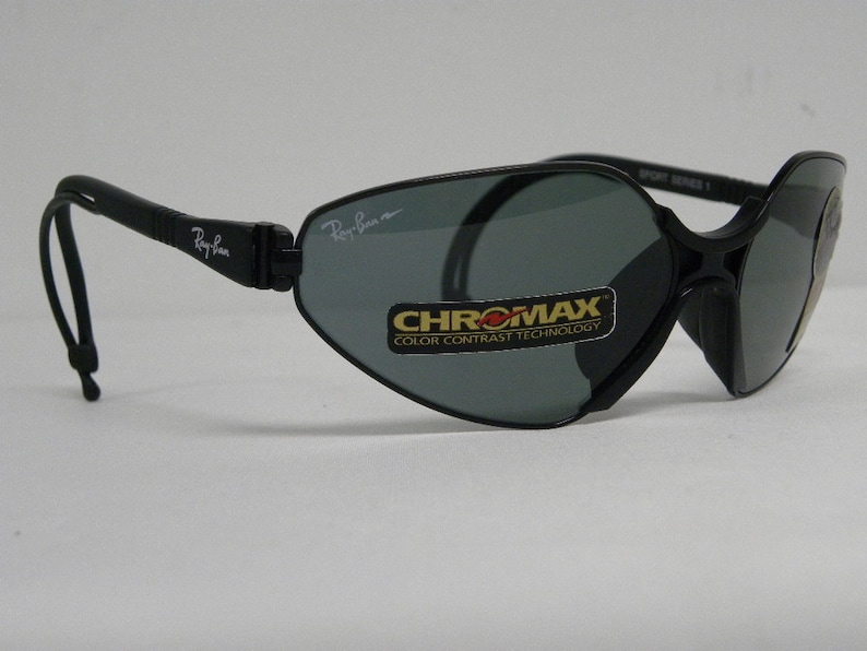 May include: Black Ray-Ban sunglasses with a gray lens and a black strap. The sunglasses have a label that says "CHROMAXX COLOR CONTRAST TECHNOLOGY" on the right lens.