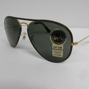 May include: Brown leather aviator sunglasses with green lenses and a Ray-Ban label. The label says "G-15 Lens" and features the Olympic rings.