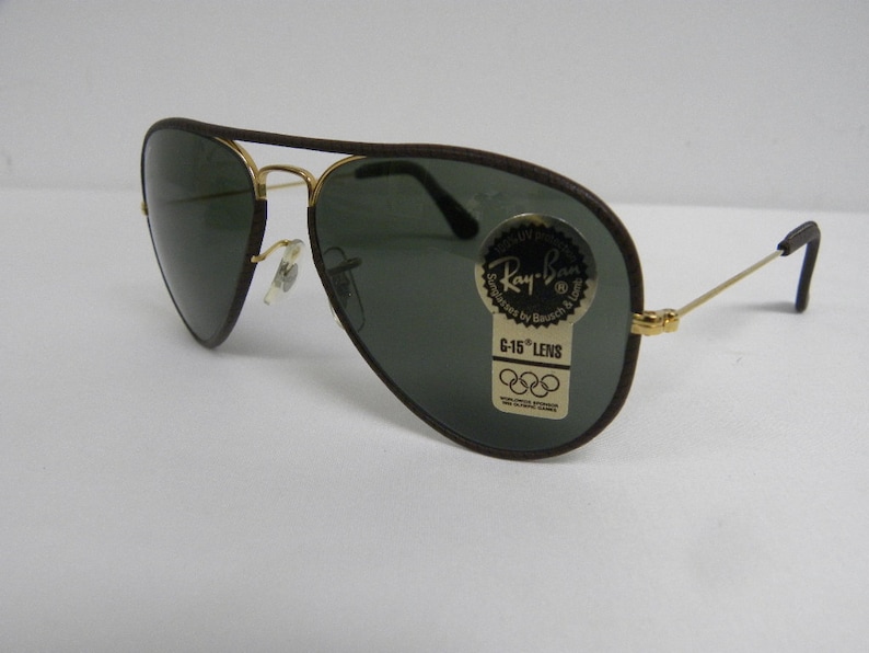 May include: Brown leather aviator sunglasses with green lenses and a Ray-Ban label. The label says "G-15 Lens" and features the Olympic rings.
