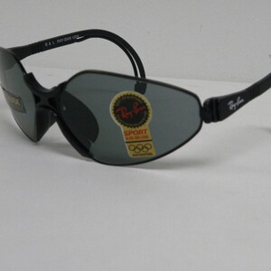 May include: Black Ray-Ban sunglasses with a gray lens and a black strap. The sunglasses have a sticker on the lens that says "Ray-Ban Sport" and has the Olympic rings logo.