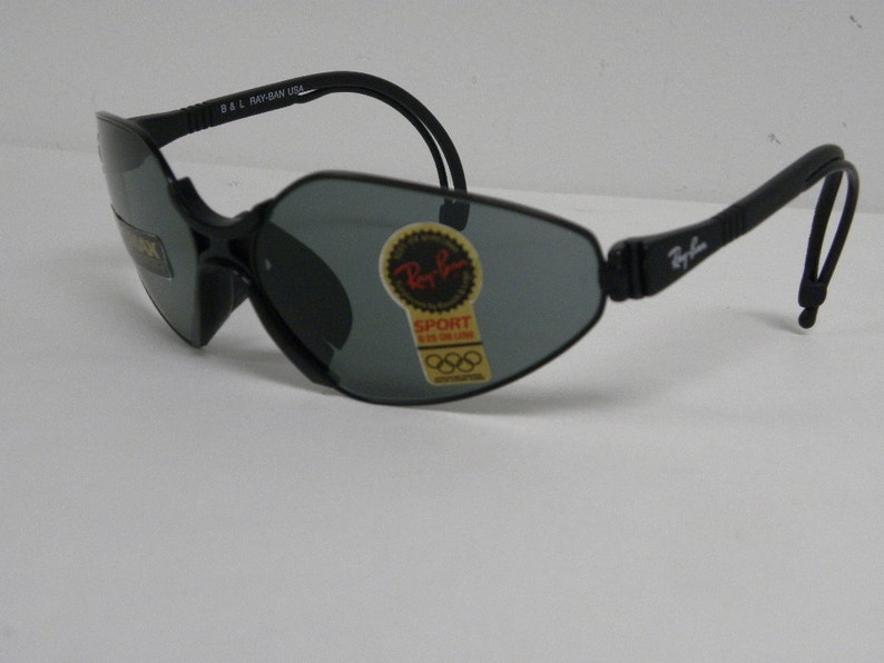 May include: Black Ray-Ban sunglasses with a gray lens and a black strap. The sunglasses have a sticker on the lens that says "Ray-Ban Sport" and has the Olympic rings logo.