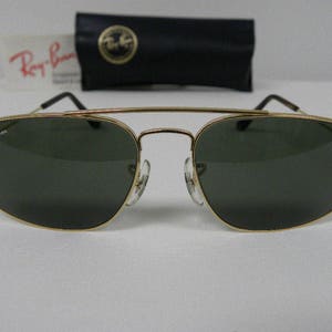 May include: Gold-framed sunglasses with dark green lenses. The Ray-Ban logo is visible on the lens and frame. Includes a black case with a gold emblem and a white card with red text.