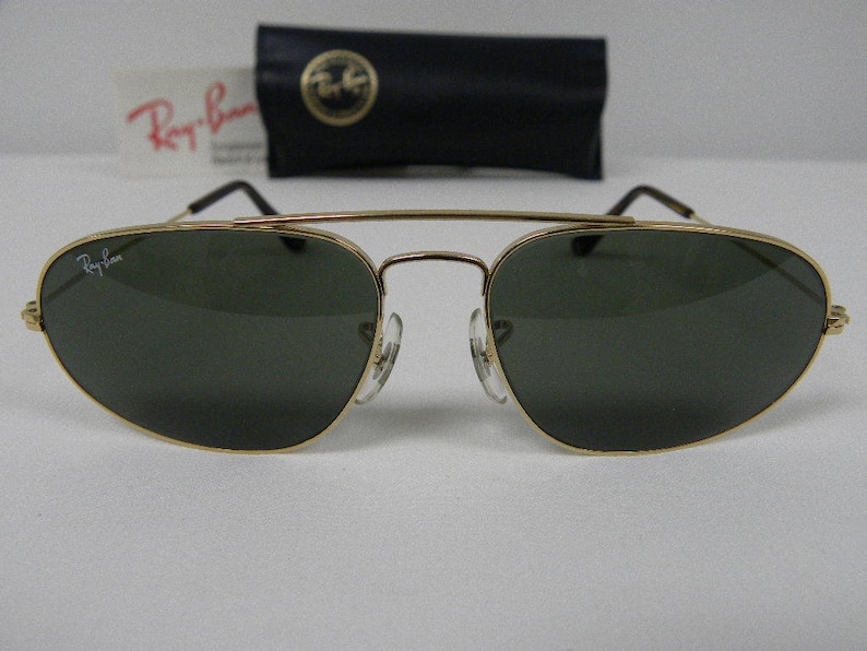 May include: Gold-framed sunglasses with dark green lenses. The Ray-Ban logo is visible on the lens and frame. Includes a black case with a gold emblem and a white card with red text.