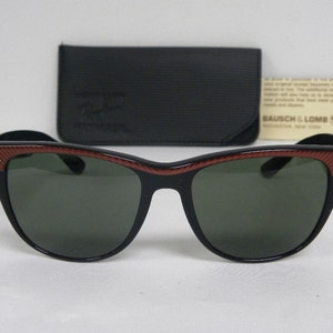 May include: Black sunglasses with red and black striped frames and green lenses. The sunglasses are sitting on a white surface. The brand name "Ray-Ban" is visible on the case.