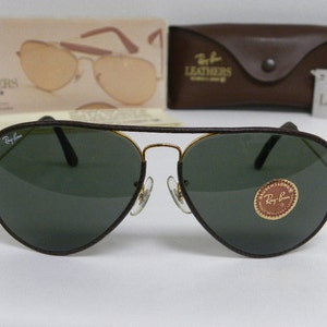 Może przedstawiać: Brązowe sk&oacute;rzane okulary przeciwsłoneczne typu aviator z zielonymi szkłami i złotymi akcentami. Okulary mają logo Ray-Ban na lewej soczewce i etykietę Ray-Ban Leather na prawej soczewce.