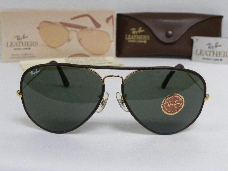Może przedstawiać: Brązowe sk&oacute;rzane okulary przeciwsłoneczne typu aviator z zielonymi szkłami i złotymi akcentami. Okulary mają logo Ray-Ban na lewej soczewce i etykietę Ray-Ban Leather na prawej soczewce.