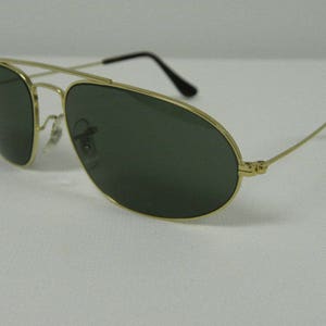 May include: Aviator-style sunglasses with a gold-colored metal frame and dark green lenses. The sunglasses have a double bridge and thin temples with black tips. The lenses are oval-shaped.