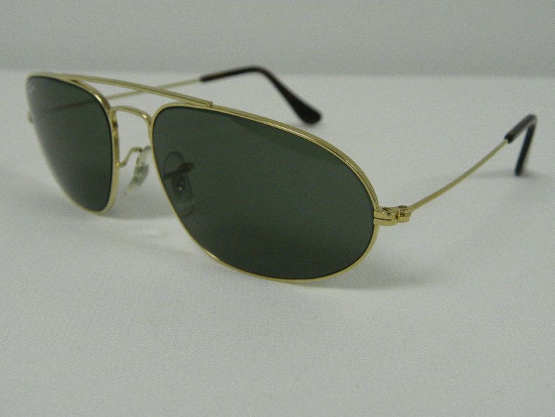 May include: Aviator-style sunglasses with a gold-colored metal frame and dark green lenses. The sunglasses have a double bridge and thin temples with black tips. The lenses are oval-shaped.