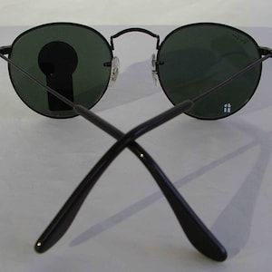 K&ouml;nnte beinhalten: Schwarze runde Sonnenbrille mit gr&uuml;nen Gl&auml;sern. Die Sonnenbrille ist zusammengeklappt, die B&uuml;gel sind gekreuzt.