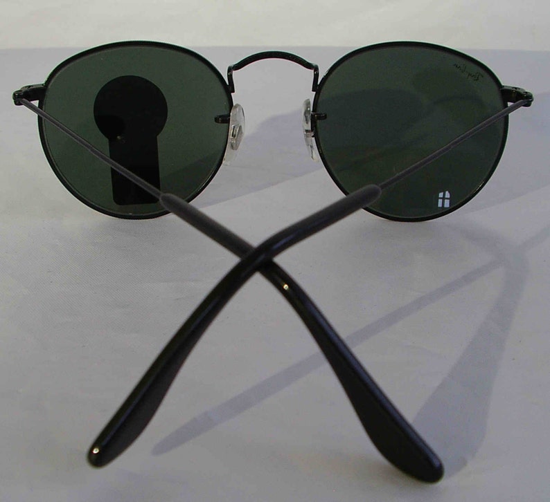 K&ouml;nnte beinhalten: Schwarze runde Sonnenbrille mit gr&uuml;nen Gl&auml;sern. Die Sonnenbrille ist zusammengeklappt, die B&uuml;gel sind gekreuzt.