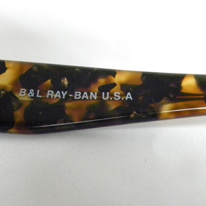 K&ouml;nnte beinhalten: Nahaufnahme des braunen Schildpatt-Rahmens einer Ray-Ban Sonnenbrille. Der Text "B&L RAY-BAN U.S.A." ist in Wei&szlig; auf den Rahmen gedruckt.