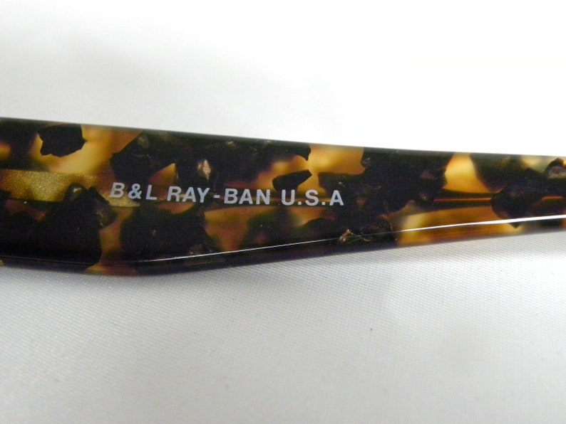 K&ouml;nnte beinhalten: Nahaufnahme des braunen Schildpatt-Rahmens einer Ray-Ban Sonnenbrille. Der Text "B&L RAY-BAN U.S.A." ist in Wei&szlig; auf den Rahmen gedruckt.