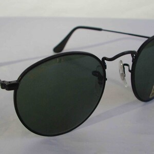 K&ouml;nnte beinhalten: Schwarze runde Sonnenbrille mit gr&uuml;nen Gl&auml;sern. Die Sonnenbrille hat einen Metallrahmen und ist neu. Die Sonnenbrille hat ein Ray-Ban-Logo auf der rechten Linse.