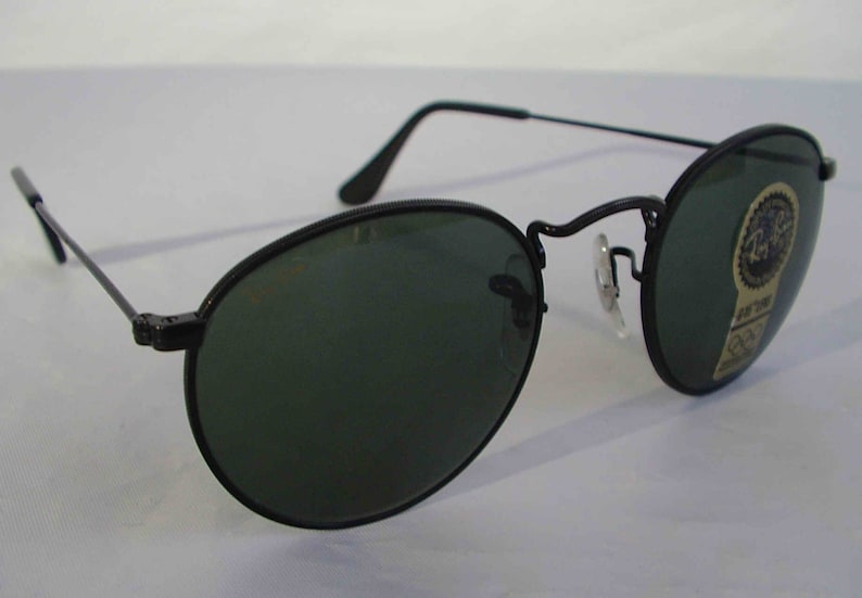 K&ouml;nnte beinhalten: Schwarze runde Sonnenbrille mit gr&uuml;nen Gl&auml;sern. Die Sonnenbrille hat einen Metallrahmen und ist neu. Die Sonnenbrille hat ein Ray-Ban-Logo auf der rechten Linse.