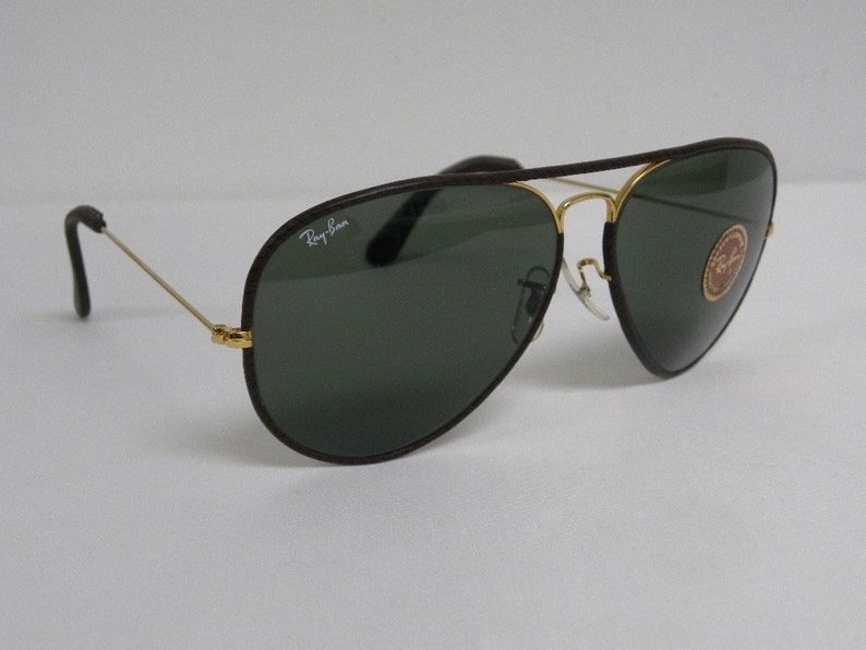 Może przedstawiać: Brązowe okulary przeciwsłoneczne typu aviator ze złotymi akcentami i zielonymi szkłami. Okulary mają logo Ray-Ban na oprawce.